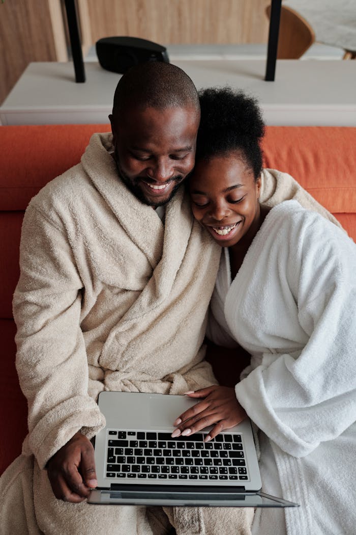 A joyful couple in bathrobes sharing a moment while browsing on a laptop indoors.